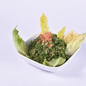 A stylish angled view of a fresh parsley tabbouleh salad in a white square bowl, topped with diced tomato, and surrounded by lemon wedges and crisp green lettuce leaves on a light purple background.