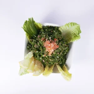 A top-down view of a fresh Tabbouleh salad in a white square bowl, topped with diced tomatoes, surrounded by green lettuce leaves and sliced lemon wedges on a white background.