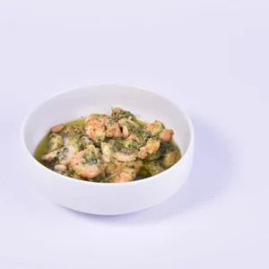 Angled close-up of a white ceramic bowl filled with premium sautéed shrimp coated in a fresh green herb, garlic, and olive oil sauce on a white background.