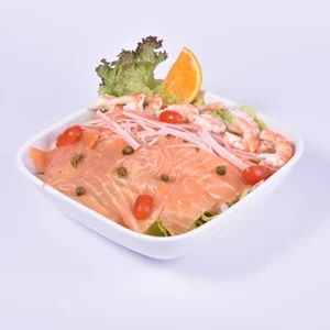 High-angle gourmet shot of a modern seafood bowl featuring layered smoked salmon, cooked prawns, cherry tomatoes, capers, onions, and an orange garnish in a white ceramic bowl.