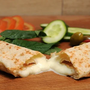 Close-up shot of a warm toasted cheese wrap torn in half, showing gooey melted white cheese stretching out, served with fresh tomato, cucumber, spinach, and olives on a wooden board.