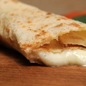 Extreme close-up shot of a toasted flour tortilla wrap broken open, revealing rich, creamy melted white cheese oozing out, resting on a wooden board.