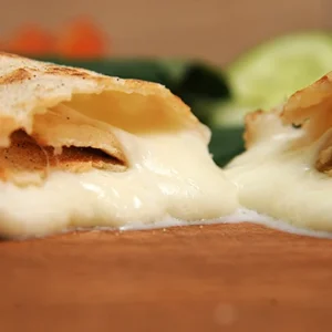 Extreme low-angle macro shot of a golden toasted tortilla wrap split in two, with rich, melted white cheese oozing out and pooling on a rustic wooden cutting board.