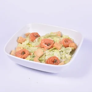Close-up angled view of a white square bowl filled with fresh Caesar salad, featuring romaine lettuce, tomato slices, golden croutons, and a creamy white dressing.