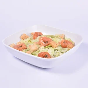 Top-down angled view of a fresh Caesar salad in a white square bowl, featuring chopped romaine lettuce, red tomato slices, and golden croutons on a white background.