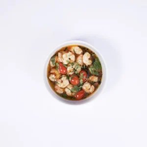 Overhead flat-lay shot of a beautiful shrimp wellness broth in a white bowl, featuring cooked shrimp, cherry tomatoes, fresh green herbs, and a clear golden broth on a white background.