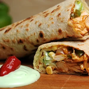 Close-up macro shot of a grilled chicken wrap cut in half, revealing grilled chicken, lettuce, corn, and bell peppers, served with a creamy green avocado dip and a red chili pepper on a wooden board.