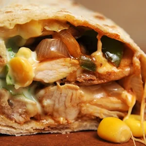 Extreme macro close-up of a grilled chicken wrap cut open, showing tender chicken, caramelized onions, bell peppers, corn, and melted white cheese oozing out on a wooden surface.
