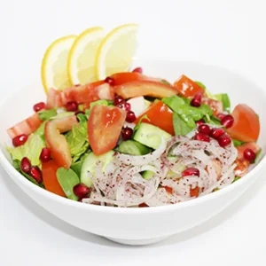 A vibrant Mediterranean salad served in a white bowl, featuring fresh greens, tomato wedges, red onion slices, pomegranate seeds, and lemon slices on a white background.