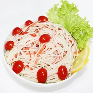 A white bowl filled with shredded crab stick salad, garnished with crisp lettuce, vibrant cherry tomatoes, and a lemon wedge on a white background.