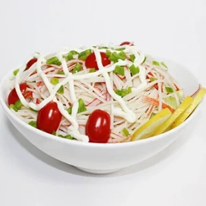 A white bowl filled with shredded crab stick salad, garnished with fresh lettuce, cherry tomatoes, white sesame seeds, and a lemon wedge on a white background .