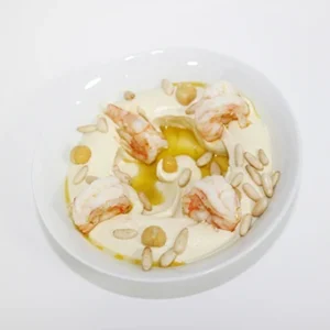 A white bowl filled with creamy hummus topped with three grilled shrimp, a swirl of olive oil, and toasted pine nuts on a clean white background.