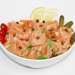 A white bowl filled with silky smoked salmon slices, garnished with green capers, pickled gherkins, cherry tomatoes, and lemon wedges.