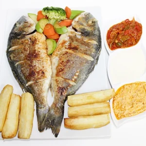 A white plate featuring two whole grilled Dorado fish, potato wedges, steamed vegetables, and three bowls of dipping sauces.
