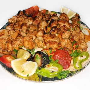 A large platter of juicy grilled shish tawook chicken chunks over flatbread, served with grilled vegetables, parsley, and lemon wedges on a silver tray.