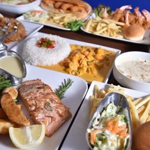 A grilled salmon fillet served with sauteed vegetables and golden potato wedges, accompanied by a creamy sauce boat, surrounded by a feast of rice, shrimp, and salads.
