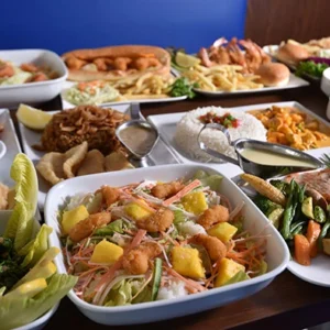 A buffet table with a large white bowl of shrimp and mango salad surrounded by grilled salmon, rice, fried shrimp, and french fries on a wooden table.