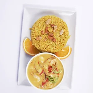 A white plate with a mound of yellow saffron rice topped with nuts, a bowl of creamy shrimp curry, and fresh orange slices on the side.