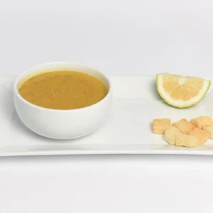 A white bowl of creamy yellow lentil soup with golden croutons and a lemon wedge on a white rectangular plate.