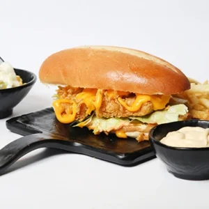 A crispy chicken cheeseburger on a black serving board, served with french fries, a bowl of coleslaw, and dipping sauce.