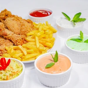 A white platter filled with golden crispy fried chicken pieces and french fries, surrounded by five small white bowls of colorful dipping sauces including red ketchup, creamy garlic, mint, and spicy sauces, garnished with fresh herbs.