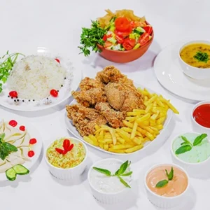 A wide top-down view of a white table featuring a large platter of crispy chicken and fries, surrounded by plates of white rice, fresh salad, hummus, lentil soup, and multiple small bowls of colorful dipping sauces.