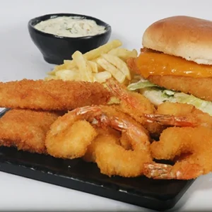 A black serving platter featuring a crispy fish sandwich with melted cheese, fried shrimp, fish fingers, and french fries, accompanied by a bowl of creamy tartar sauce.