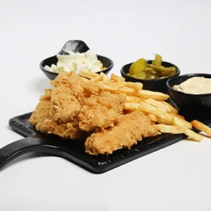 A black serving board with a handle featuring golden crispy chicken tenders and french fries, surrounded by three black bowls containing creamy coleslaw, green pickles, and garlic dip.