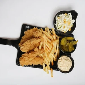 A black serving board featuring golden crispy chicken tenders and french fries, accompanied by small black bowls of creamy coleslaw, pickles, and dipping sauce.