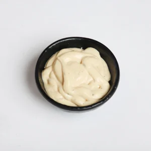 A small black bowl filled with a thick, creamy white garlic sauce with a smooth swirl pattern on the surface, set against a plain white background.