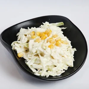 A black leaf-shaped bowl filled with a creamy shredded cabbage slaw, topped with yellow sweet corn kernels, set against a bright white background.