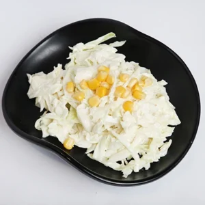 A top-down view of a black wavy plate filled with freshly shredded white cabbage, topped with scattered yellow sweet corn kernels, set against a plain white background.