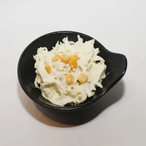 A close-up of a black leaf-shaped bowl filled with creamy white shredded coleslaw, generously topped with bright yellow sweet corn kernels, set against a plain white background.