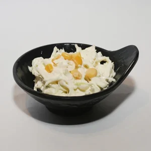 A close-up of a black leaf-shaped bowl filled with creamy white coleslaw, topped with bright golden yellow sweet corn kernels, set against a plain light grey background.