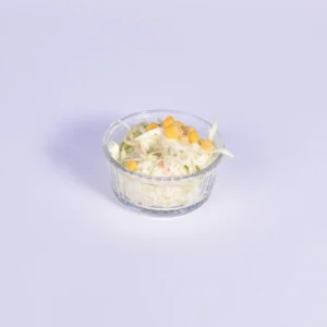 A close-up shot of a small clear glass bowl filled with creamy white shredded coleslaw, topped with bright yellow sweet corn kernels, placed on a light lavender-white background.