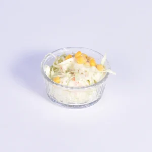 A small clear glass bowl filled with creamy white shredded cabbage coleslaw, topped with yellow sweet corn kernels, set against a plain white background.