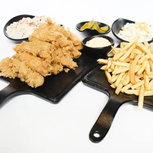 A perspective view of two black slate serving boards; the left board holds crispy fried chicken tenders with a small bowl of white dip, the right board holds a large pile of golden french fries. Behind the boards are three black bowls containing spiced white rice, green pickles, and creamy coleslaw.