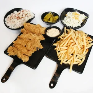 A restaurant-style spread featuring black serving boards with crispy fried chicken strips and a large portion of golden french fries, surrounded by black bowls of white rice, creamy coleslaw, green pickles, and a small cup of dipping sauce, set against a white background.