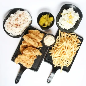 A top-down view of a black serving platter set featuring crispy fried chicken strips, a bowl of white rice, a pile of golden french fries, a bowl of creamy coleslaw, a bowl of green pickles, and a small cup of white dipping sauce.