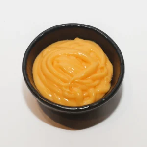 A close-up top-down view of a small black bowl filled with a thick, glossy, and swirled creamy orange sauce, set against a plain white background.