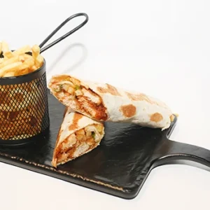 A black serving board presenting a grilled shawarma wrap cut into two halves revealing a juicy meat filling, accompanied by a black metal basket filled with golden crispy french fries.