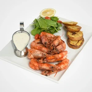 A square white platter featuring a stack of grilled whole shrimp, a metal gravy boat with creamy white sauce, roasted potato wedges, fresh green salad with cherry tomatoes, and a lemon slice on top.